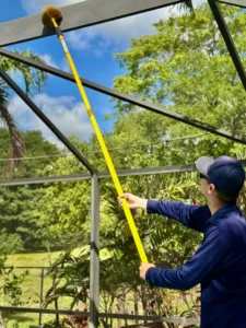 Pest control technician cleaning pool cage screen enclosure with long-handled brush in South Florida backyard