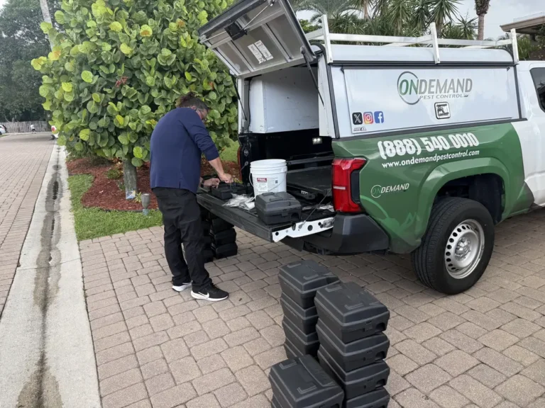 On Demand Pest Control technician loading black rodent bait stations from a branded service truck bed